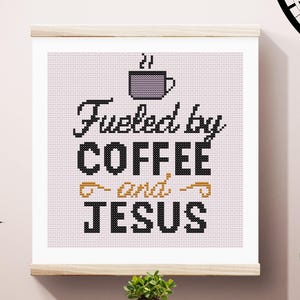 May include: Cross-stitch art framed in a light wood hanger. The design features the phrase "Fueled by COFFEE and JESUS" in black and gold lettering, with a gray coffee cup icon above the word "COFFEE". The background is a light pink grid.