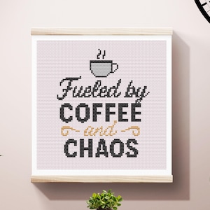 May include: Cross-stitch artwork in a light wood frame, featuring the phrase "Fueled by COFFEE and CHAOS" in black and gold lettering, with a coffee cup icon. The artwork is displayed on a light pink background.