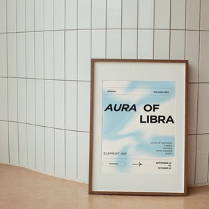 May include: A framed poster with a brown wooden frame. The poster features the text "AURA OF LIBRA" in bold black letters against a light blue and white gradient background. Additional text includes "ELEMENT AIR" and dates.