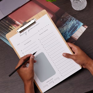 May include: A wooden clipboard with a daily planner sheet. The sheet has sections for top priorities, a daily routine, and notes. A black pen is held over the notes section. The words "daily planner" are written at the top.