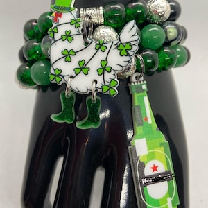 Könnte beinhalten: Ein grünes und weißes Armband mit einem grünen Kleeblatt und einem grünen Bierflaschen-Anhänger. Das Armband besteht aus grünen Perlen und hat einen silbernen Verschluss.