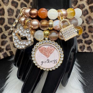 May include: A collection of beaded bracelets in shades of gold, brown, and white. The bracelets feature charms including a rhinestone-studded padlock, lips, and a pendant with the text "girlboss". The background has a leopard print.