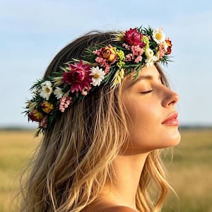 Può includere: Una corona di fiori composta da un mix di fiori secchi, tra cui elicrisi color magenta intenso, piccole margherite bianche, fiori rosa di riempimento e fogliame verde. La composizione è circolare e pensata come accessorio per capelli.