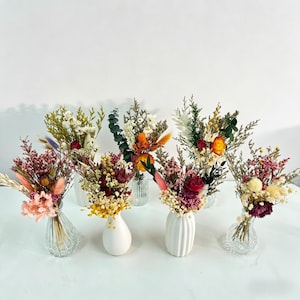 May include: A collection of dried flower bouquets in various colors, including pink, yellow, and orange. The bouquets are arranged in small glass and white ceramic vases, creating a rustic and natural aesthetic.