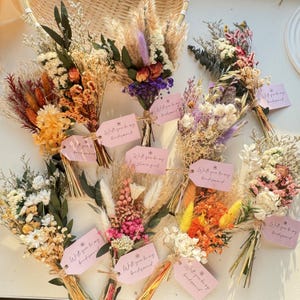 May include: A collection of dried flower bouquets in various colors, including yellow, orange, purple, and white. Each bouquet is tied with twine and features a pink tag with the text "Will you be my bridesmaid" or "Will you be my flower girl".