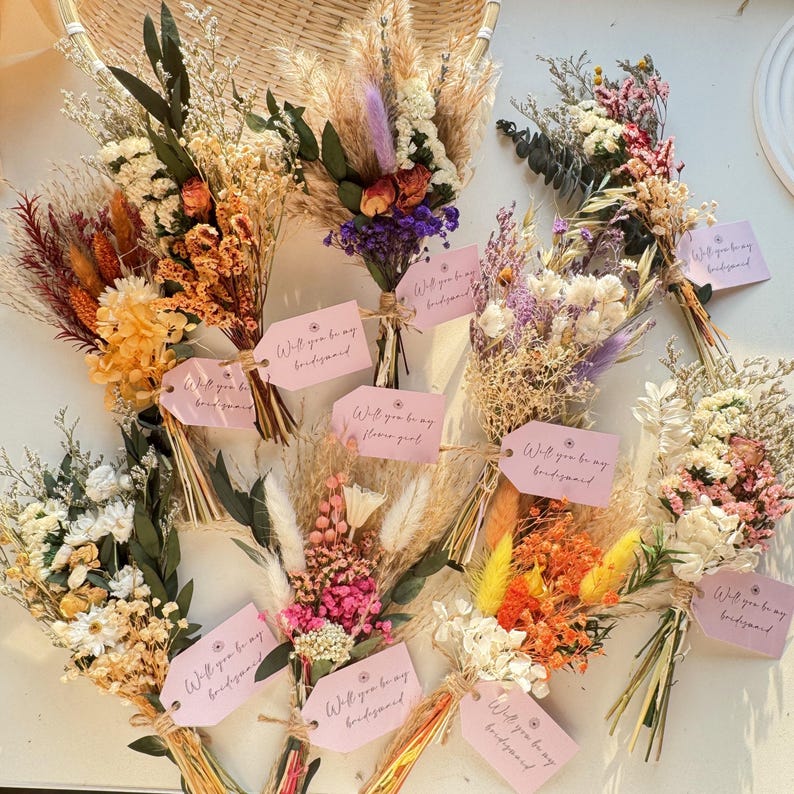 May include: A collection of dried flower bouquets in various colors, including yellow, orange, purple, and white. Each bouquet is tied with twine and features a pink tag with the text "Will you be my bridesmaid" or "Will you be my flower girl".