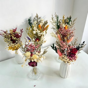 May include: Four small glass and ceramic vases containing arrangements of dried flowers in various colors including pink, yellow, white, and deep red. The bouquets feature diverse textures with sprigs of greenery, fluffy grasses, and small blossoms.