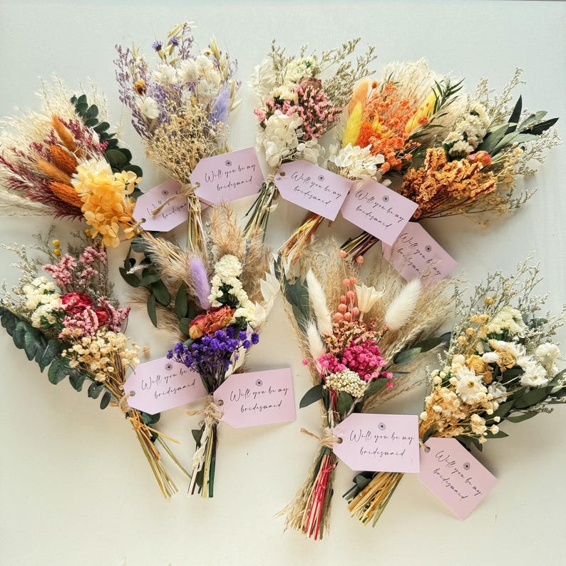 May include: An assortment of dried flower bouquets in various colors, including orange, pink, and purple. Each bouquet is tied with twine and features a pink tag that reads "Will you be my bridesmaid".