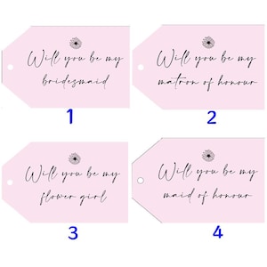 May include: Four pink gift tags with the text "Will you be my bridesmaid", "Will you be my matron of honor", "Will you be my flower girl", and "Will you be my maid of honor". Each tag has a small daisy graphic.