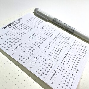 K&ouml;nnte beinhalten: Ein wei&szlig;er Kalender f&uuml;r 2021 mit monatlichen Layouts, der ein minimalistisches Design und handgeschriebene Monatsnamen aufweist. Ein grauer Stift liegt in der oberen rechten Ecke des Kalenders, der auf einer gepunkteten Oberfl&auml;che liegt.