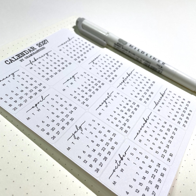 K&ouml;nnte beinhalten: Ein wei&szlig;er Kalender f&uuml;r 2021 mit monatlichen Layouts, der ein minimalistisches Design und handgeschriebene Monatsnamen aufweist. Ein grauer Stift liegt in der oberen rechten Ecke des Kalenders, der auf einer gepunkteten Oberfl&auml;che liegt.