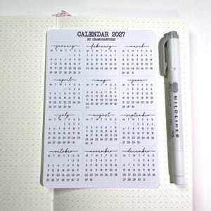 K&ouml;nnte beinhalten: Ein wei&szlig;es Aufkleberblatt mit einem Kalenderdesign f&uuml;r 2027, mit Monaten in Handschrift. Ein grauer Mildliner Textmarker befindet sich neben dem Kalender. Der Kalender befindet sich auf einem karierten Papier.