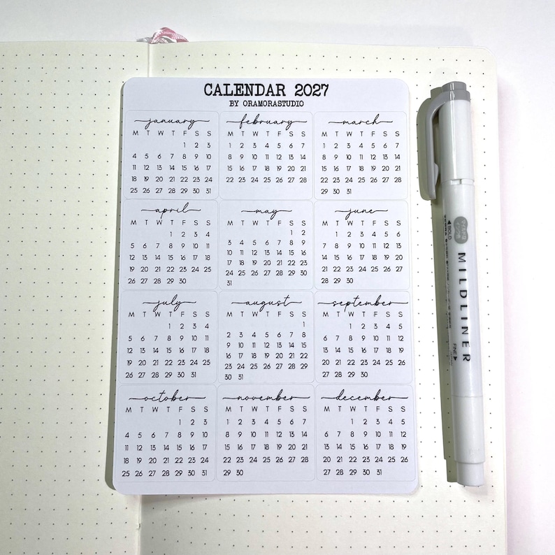 K&ouml;nnte beinhalten: Ein wei&szlig;es Aufkleberblatt mit einem Kalenderdesign f&uuml;r 2027, mit Monaten in Handschrift. Ein grauer Mildliner Textmarker befindet sich neben dem Kalender. Der Kalender befindet sich auf einem karierten Papier.