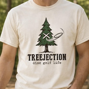 May include: An ivory short-sleeve t-shirt featuring a graphic of a green pine tree with a white X and a flying disc. Below the tree, the text reads TREEJECTION disc golf life in a dark brown font. The shirt has a crew neckline and a standard fit.