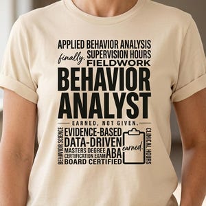 Pode incluir: Camiseta creme com as palavras "BEHAVIOR ANALYST" em letras pretas grandes. Texto adicional inclui "APPLIED BEHAVIOR ANALYSIS", "SUPERVISION HOURS" e "FIELDWORK". A camiseta também lista "EVIDENCE-BASED" e "DATA-DRIVEN".