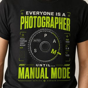 Könnte beinhalten: Ein schwarzes Kurzarm-T-Shirt mit einer Grafik eines Kameramodus-Wahlrads in der Mitte. Der Text "EVERYONE IS A PHOTOGRAPHER UNTIL... MANUAL MODE" ist in Limettengrün und Weiß gedruckt, ergänzt durch Kameraeinstellungen wie "ISO 100" und "F/2.8".
