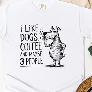 Op de afbeelding: Wit T-shirt met een zwarte afbeelding van een hond die koffie drinkt, en de tekst "I LIKE DOGS, COFFEE AND MAYBE 3 PEOPLE." Een casual shirt voor honden- en koffieliefhebbers.