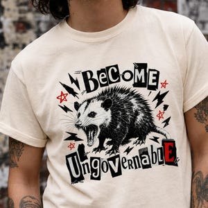 Peut inclure: Un t-shirt ivoire à manches courtes avec une illustration en noir et blanc d'un opossum hargneux. Le texte "BECOME UNGOVERNABLE" est imprimé dans une police grasse et usée, agrémenté de petites étoiles rouges et d'éclairs noirs.