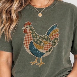 May include: A moss green short-sleeve t-shirt featuring a central graphic of a chicken composed of various patchwork patterns including red and white gingham, blue with white dots, and floral prints in shades of yellow, blue, and green.