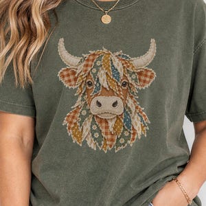 May include: A moss green short-sleeve t-shirt featuring a graphic of a highland cow. The cow illustration is composed of various patterned sections in shades of orange, blue, and white. The shirt has a crew neckline and a relaxed fit.