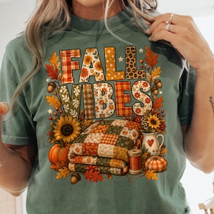 Op de afbeelding: Een mosgroen t-shirt met een grafische print met de tekst FALL VIBES in een mix van ruit-, bloemen- en luipaardpatronen. Het ontwerp bevat een gevouwen quilt, zonnebloemen, pompoenen, eikels, herfstbladeren en een kleine kan.