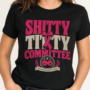 Op de afbeelding: Een zwart T-shirt met korte mouwen met de tekst SHITTY TITTY COMMITTEE in roze en beige, een roze lintafbeelding en een cirkelvormig embleem met de tekst MEMBER SINCE 2026. Het ontwerp bevat kleine hartjes en decoratieve laurierblaadjes.