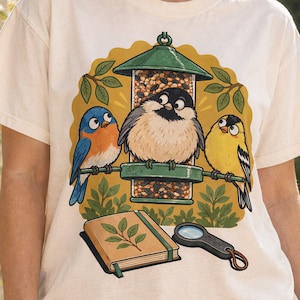 Könnte beinhalten: Ein elfenbeinfarbenes T-Shirt mit einer grafischen Illustration von drei Vögeln, die auf einem grünen, mit Samen gefüllten Vogelfutterhaus sitzen. Unter dem Futterhaus liegt ein geschlossenes beiges Notizbuch mit einem grünen Blattmotiv neben einer schwarzen Lupe.