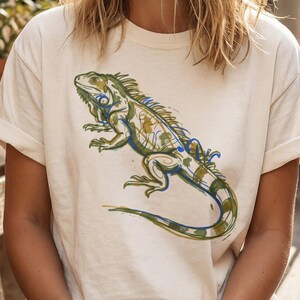 Op de afbeelding: Ivoor kleurig T-shirt met een gedetailleerde illustratie van een leguaan in groen, blauw en geel. Het leguaanontwerp is gecentreerd op de voorkant. Het shirt heeft korte mouwen en een relaxte pasvorm.