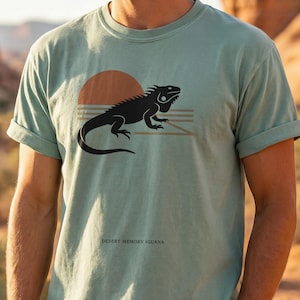 Op de afbeelding: Lichtgroen t-shirt met een zwart leguaanontwerp, een ondergaande zon en horizontale lijnen. De tekst "Desert Memory Iguana" is eronder gedrukt. De mouwen zijn opgerold.