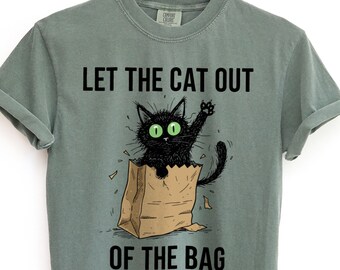May include: Moss green t-shirt with the phrase "LET THE CAT OUT OF THE BAG" printed in black. The graphic features a black cat with green eyes popping out of a brown paper bag. The shirt is hanging on a wooden hanger.