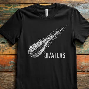 May include: Black t-shirt with a white graphic depicting a comet or space vehicle and the text "31/ATLAS". The design includes a detailed illustration of the vehicle's front. The shirt is made of a soft material.