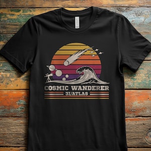 Può includere: T-shirt nero con una grafica raffigurante un tramonto, una cometa e un'onda. Il testo "COSMIC WANDERER 31/ATLAS" è stampato sotto l'onda in stile retrò. La maglietta è realizzata in materiale morbido.