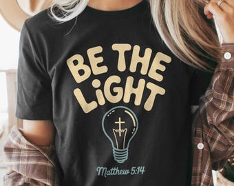 May include: Black t-shirt with the text "BE THE LIGHT" in a light yellow, bubble-style font. Below the text is a light blue lightbulb graphic with a cross inside. The text "Matthew 5:14" is below the lightbulb.
