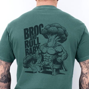 Puede incluir: Una camiseta verde con un gráfico de un superhéroe de brócoli levantando pesas. El texto "BROC AND ROLL BABY" está impreso en la camiseta.