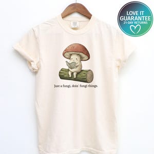 May include: An ivory short-sleeve t-shirt featuring a graphic of a cartoon mushroom sitting on a log and reading a book. Below the illustration, the text reads, "Just a fungi, doin' fungi things." The shirt is displayed on a wooden hanger.