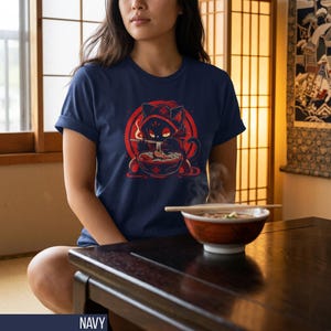 May include: Navy blue t-shirt featuring a red and black cartoon cat enjoying ramen. The cat is wearing a hood and is set against a red circle. The shirt is a classic crew neck style.