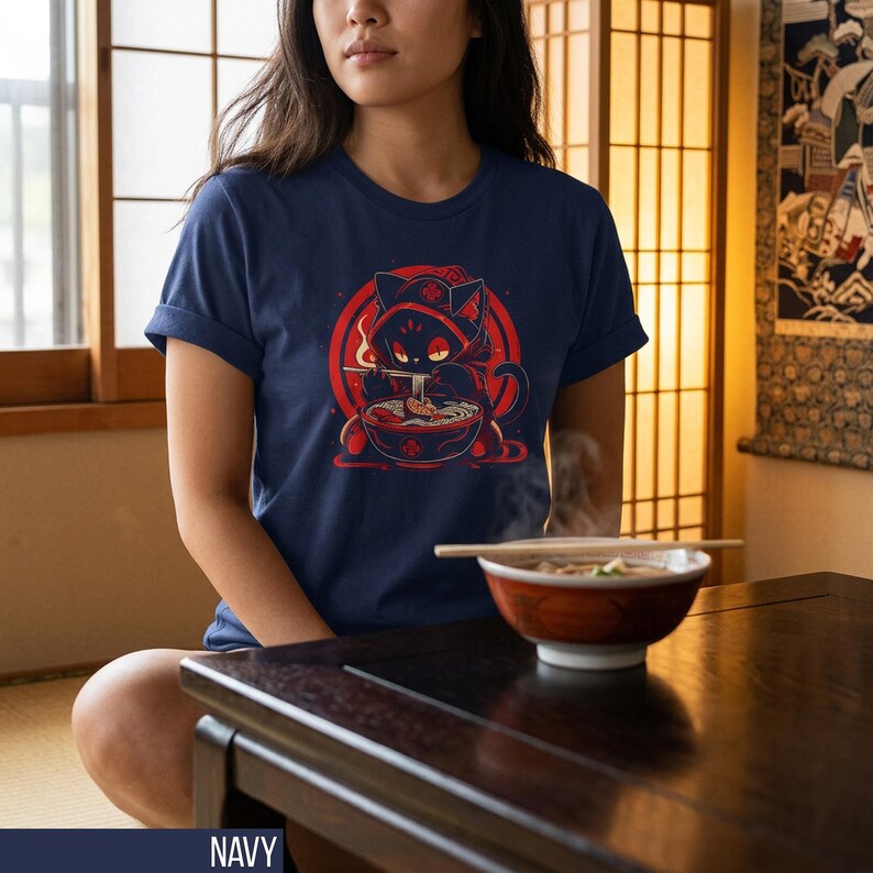 May include: Navy blue t-shirt featuring a red and black cartoon cat enjoying ramen. The cat is wearing a hood and is set against a red circle. The shirt is a classic crew neck style.