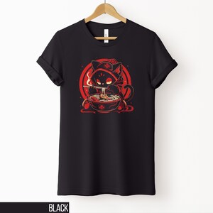 May include: Black t-shirt featuring a cartoon cat in a red hooded cloak, eating ramen from a bowl. The graphic is set against a red circular background. The word "BLACK" is printed at the bottom of the shirt.