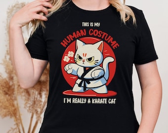 May include: Black t-shirt featuring a cartoon karate cat design. The cat is white with a black belt and a stern expression, set against a red circle. Text reads "THIS IS MY HUMAN COSTUME, I'M REALLY A KARATE CAT."
