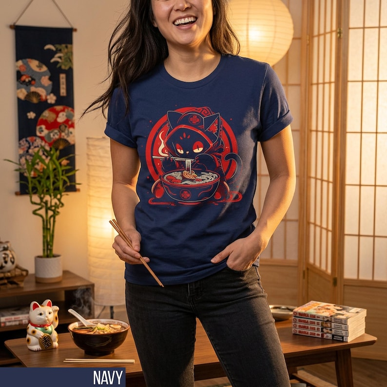 May include: Navy blue t-shirt featuring a red and black graphic of a cat eating ramen. The cat is wearing a hood and is surrounded by a red circle. The shirt is a crew neck style.