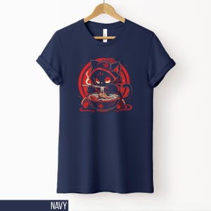 May include: Navy blue t-shirt featuring a cartoon cat in a red hooded cloak, eating ramen from a bowl. The cat is set against a red circle with a white background. The word "NAVY" is at the bottom.