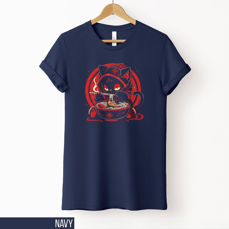 May include: Navy blue t-shirt featuring a cartoon cat in a red hooded cloak, eating ramen from a bowl. The cat is set against a red circle with a white background. The word "NAVY" is at the bottom.