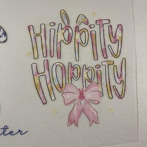 May include: A pastel-colored graphic with the words "Hippity Hoppity" in a whimsical font, outlined in black. A pink bow with a bunny face is centered below the text. Easter-themed design with star accents.