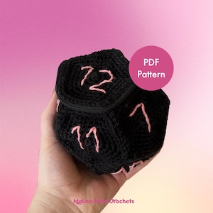 Op de afbeelding: Een zwarte gehaakte dobbelsteentas met een ritssluiting en roze geborduurde nummers. De tas wordt in een hand gehouden. De tekst "PDF Pattern" staat in een roze cirkel.