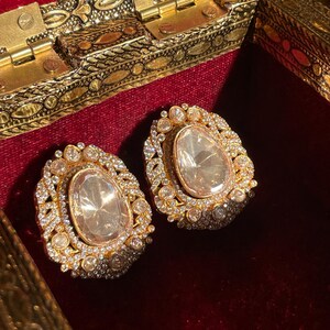 May include: A pair of gold earrings featuring large, oval, clear centre stones. The earrings are encircled by smaller, clear stones and detailed goldwork. They sit on a red velvet surface within a decorative gold box.