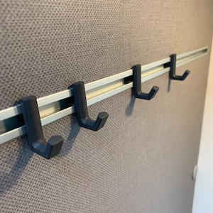 Könnte beinhalten: Vier schwarze Metallhaken, die an einer silbernen Schiene befestigt sind, die an einer strukturierten grauen Wand befestigt ist.