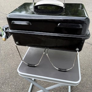 Könnte beinhalten: Ein schwarzer tragbarer Grill mit silbernem Griff und Beinen. Der Grill steht auf einem grauen Klapptisch mit silbernen Beinen. Der Griff besteht aus schwarzem Kunststoff.