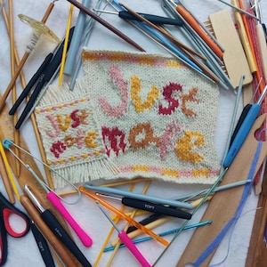 Peut inclure: Un gros plan sur un tissu tricoté avec les mots "Just Make" en fil rose, jaune et orange. Le tissu est entouré d'une variété d'aiguilles à tricoter et de crochets.
