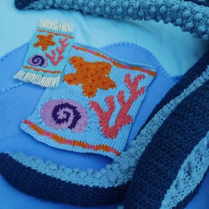 May include: A blue knitted octopus with a blue, orange, and pink knitted rug with a starfish, seashell, and coral design. The rug has a fringe border.