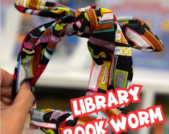 May include: A black headband with a colorful book print design. The headband is tied in a knot and the words "LIBRARY BOOK WORM" are printed in red on the front.